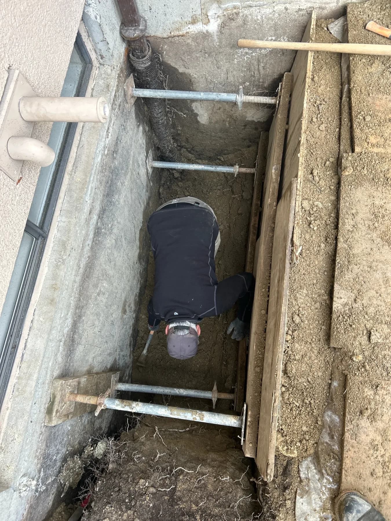 Technician working in a shored exterior drainage excavation beside a building foundation