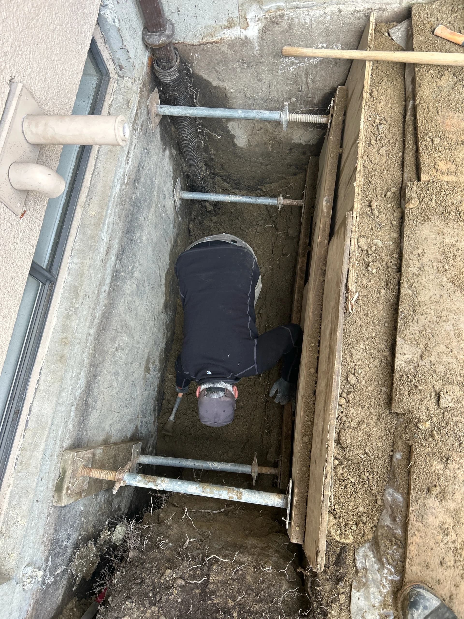 Technician working in a shored exterior drainage excavation beside a building foundation